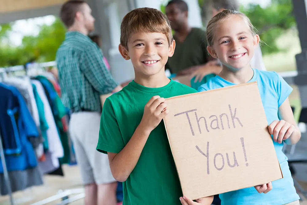 Kids holding up a sign that says 'Thank You' in front of a community event, representing gratitude for donations and sponsorships